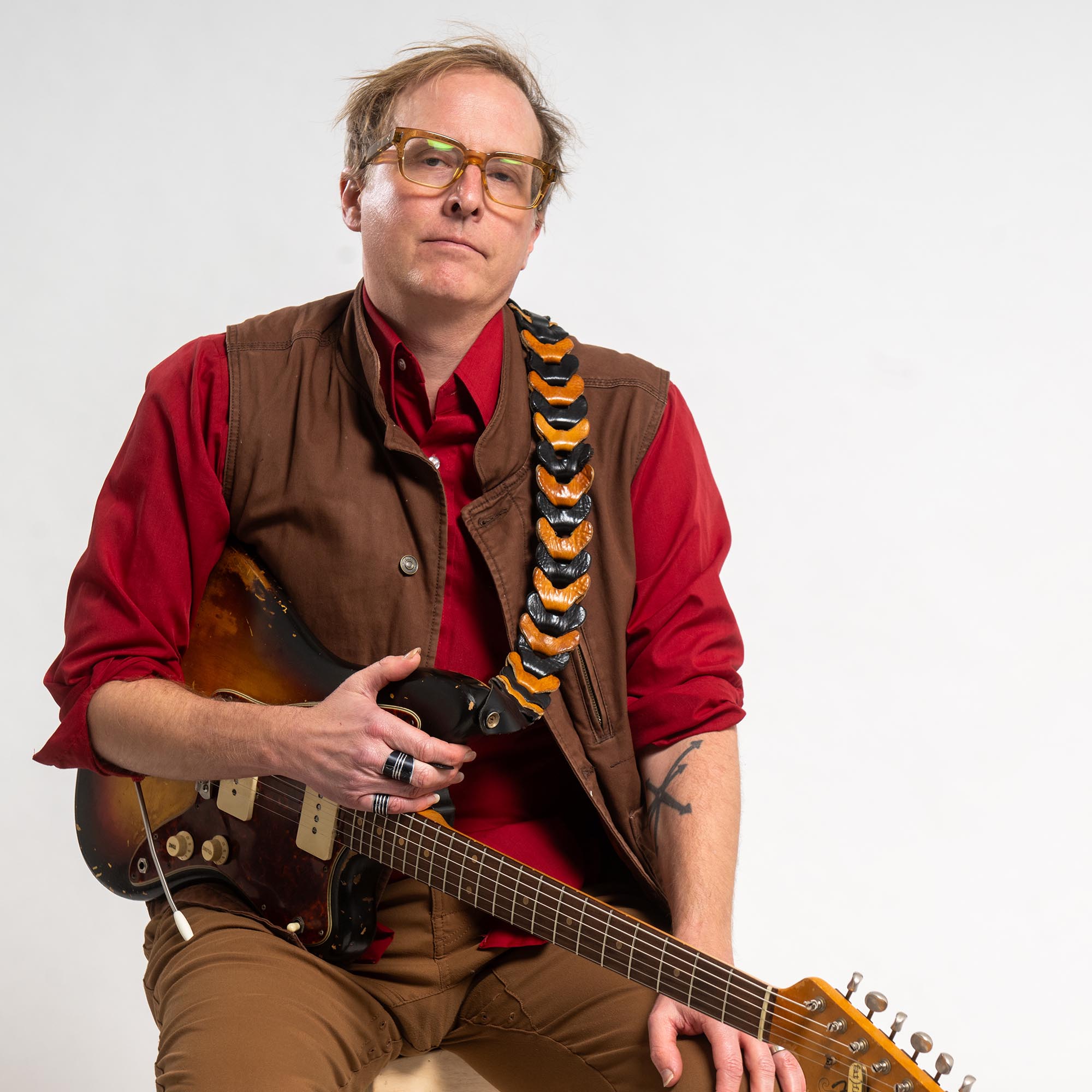 Photo: Nathaniel Braddock, a white man wearing glasses, a red collared shirt with the sleeves rolled up, a brown vest, and brown pants, sits casually in a chair and poses with his guitar in front of a white background.