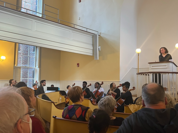 A woman with curly hair and tanned skin wearing a dark t-shirt stands at the altar of a church in front of a podium, addressing a crowd of sitting in the pews. A quartet of young Black musicians hold cellos and prepare to play. 