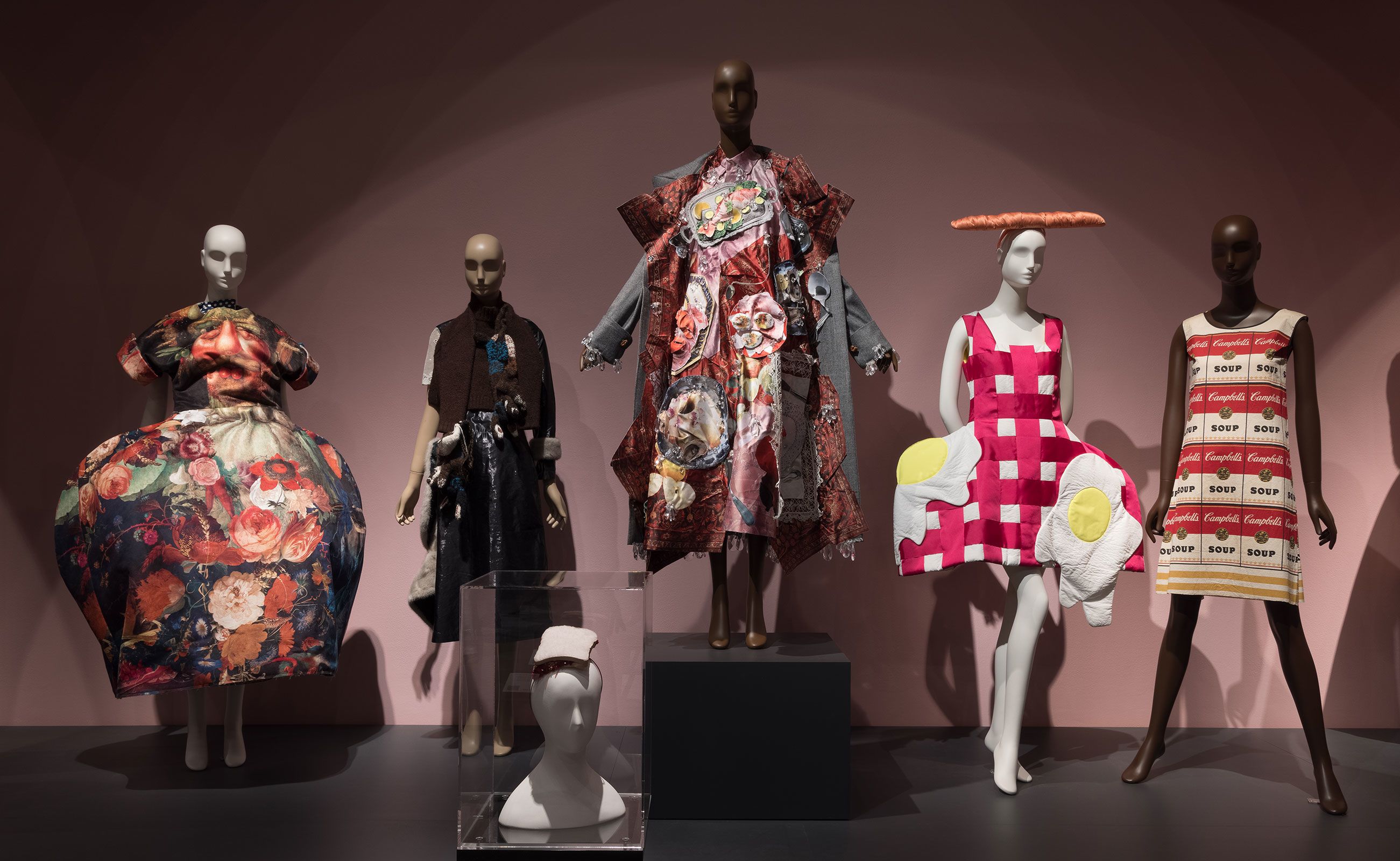 Pieces included in the exhibition's "Feeding the Eye" display include a sculptural gown from Comme des Garçons Spring-Summer 2018 collection (far left), a PB&J sandwich hat designed by Stephen Jones (foreground center) and the paper "Souper Dress" designed by Campbell's Soup Company circa-1966 (far right).