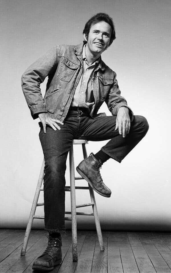 A black-and-white photo of a young Mr. Del Tredici, seated on a tall stool with his left leg bent and his left hand resting on his thigh. He is casually dressed in a jean jacket, jeans and boots, and he is smiling.