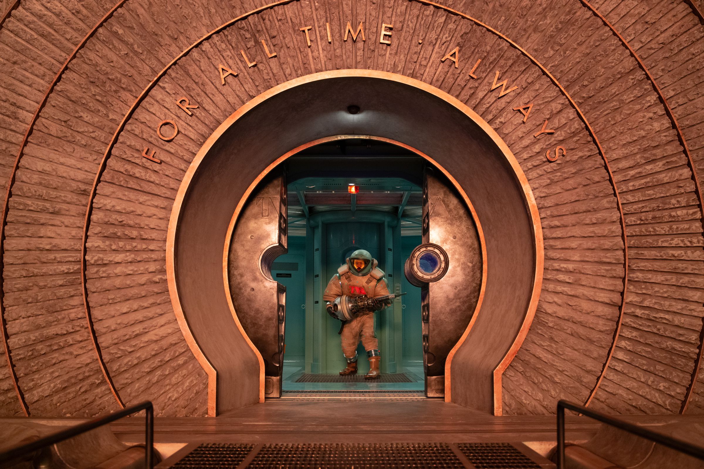 A man in a space suit holding a device that looks like a massive screw in both hands. The man is standing in the circular doorway opening onto a platform with a grated gangway. Above the door the words “for all time. Always” are inscribed.