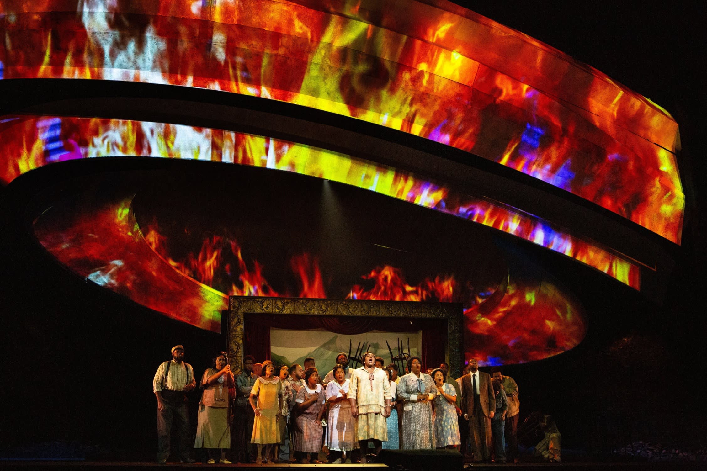 A group of people stand on a stage, singing; above them, images of multicoloured flames are projected on to a large circular structure