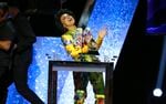 A woman in a colorful outfit stands behind a podium with a bit smile on her face and gestures enthusiastically with her hands.