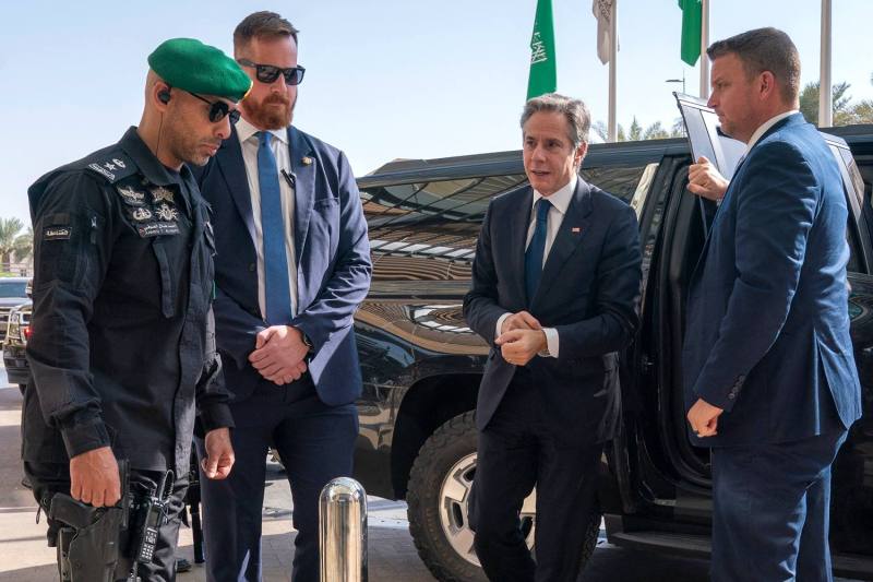 Blinken stands near a black SUV among U.S. and Saudi security personnel. A Saudi flag can be seen in the background.