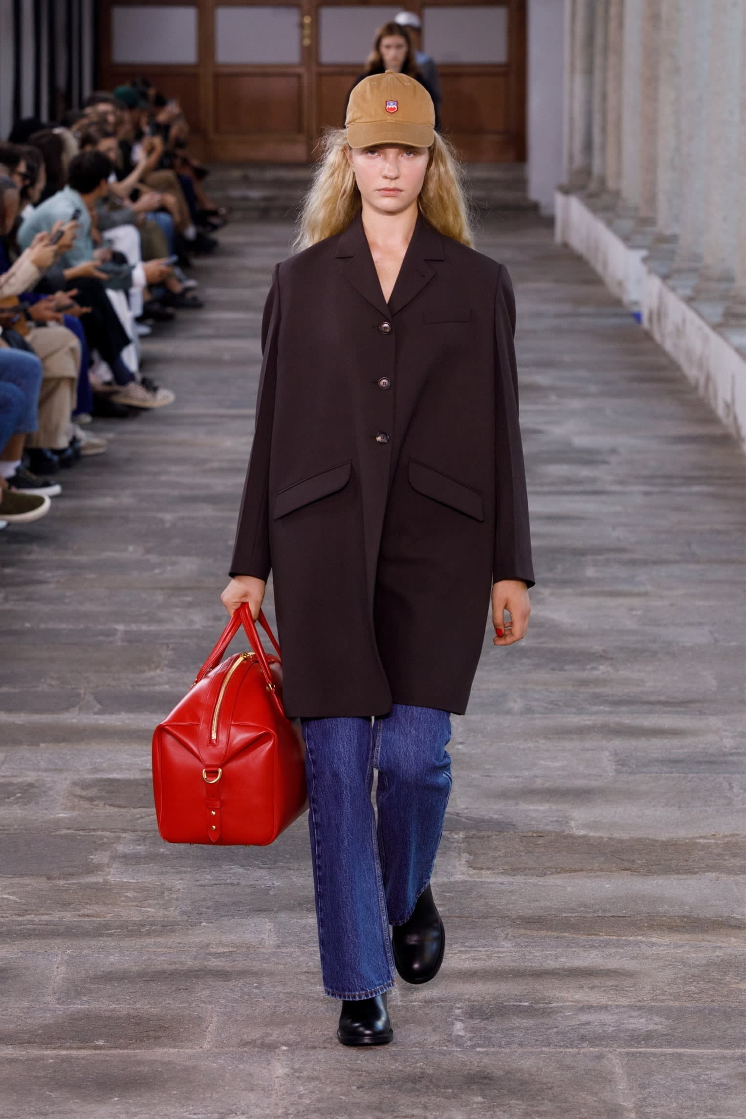 female model on runway in black overcoat, blue flared trousers and a brown baseball cap