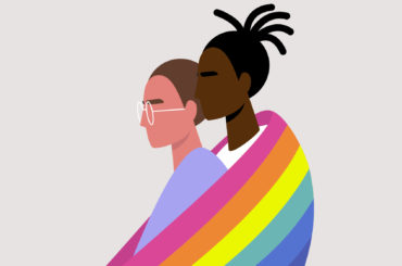 Two people hold each other as a rainbow flag wraps around them.