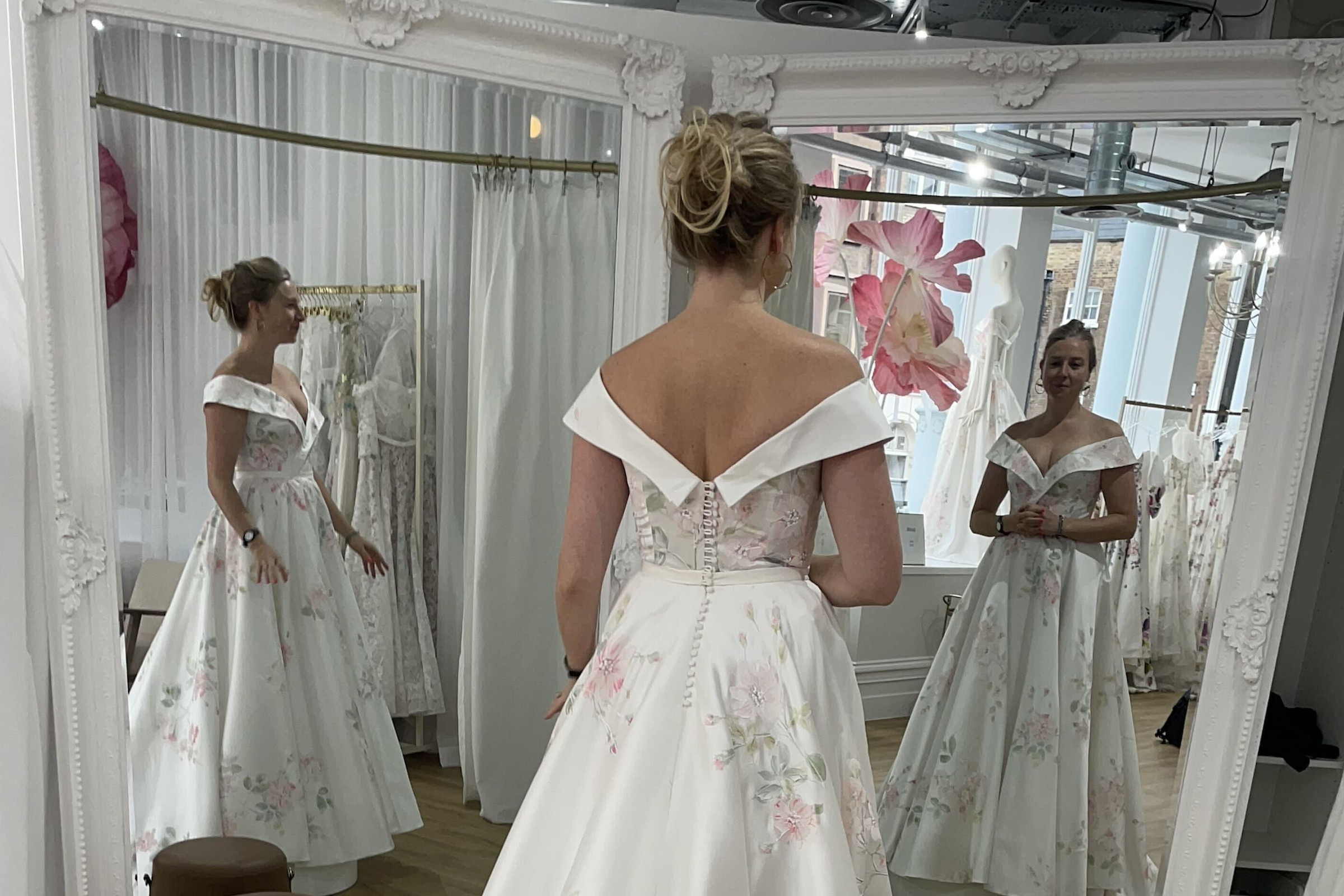 A picture of a woman, taken from behind, who is standing in a bridal gown in front of two mirrors. In all three instances of her, her hands are in different positions.