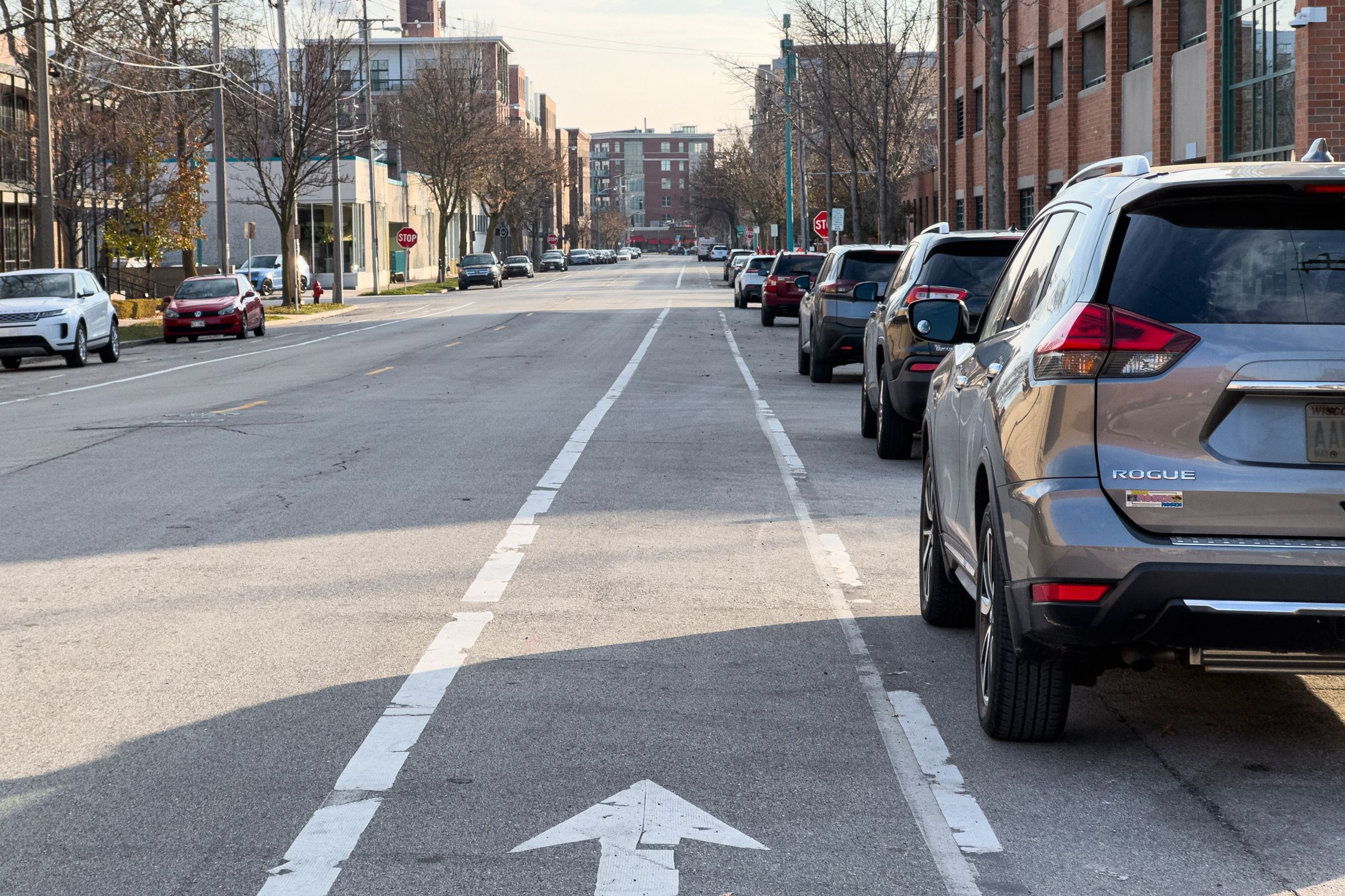 A late-in-the-day view down a bike lane that sits between a parking lane and a driving lane. Several commercial buildings and parked cars line the otherwise empty street.