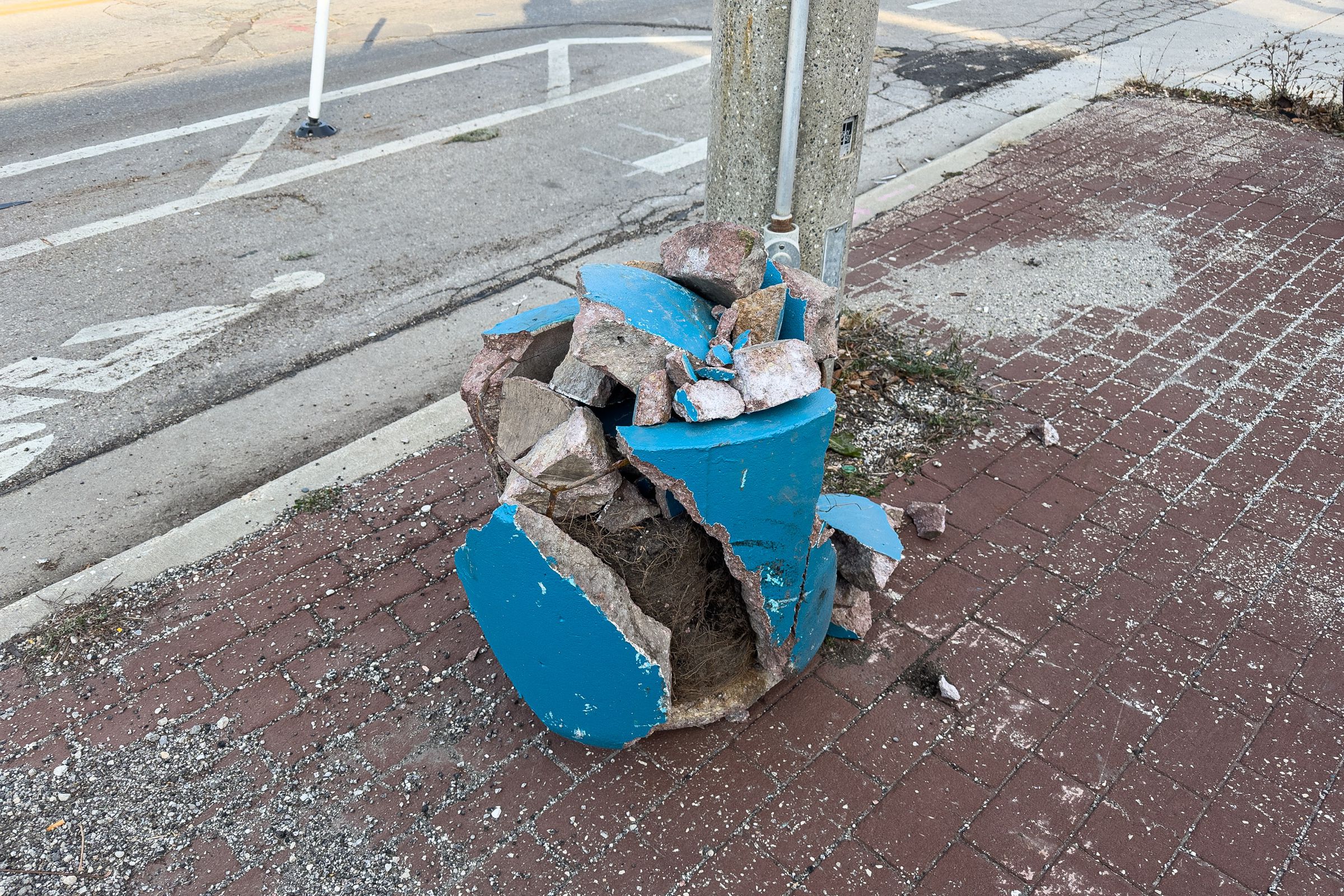 A picture of a broken planter sitting on a brick sidewalk next to a curb. Beyond that, a buffered bike lane.