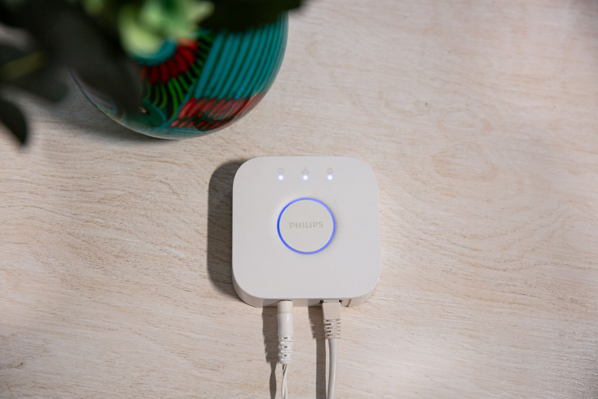 A Philips Hue Bridge on a white table with a power cord and ethernet cable connected to it.