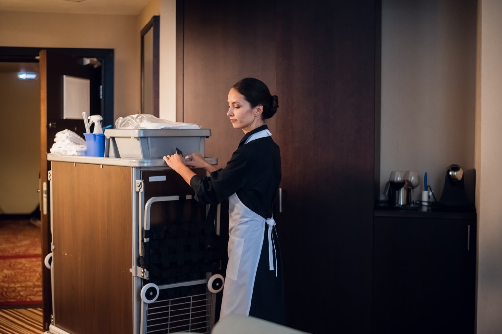 A woman in a black and white uniform pushes a housekeeping cart at a hotel. 