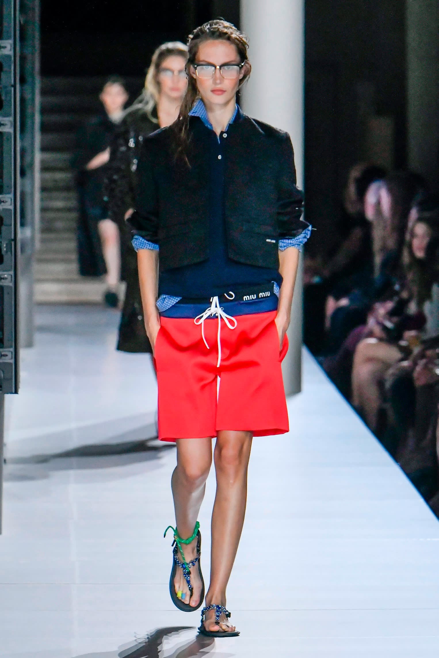 Models on a catwalk. The model at the front wears a red skirt and navy top