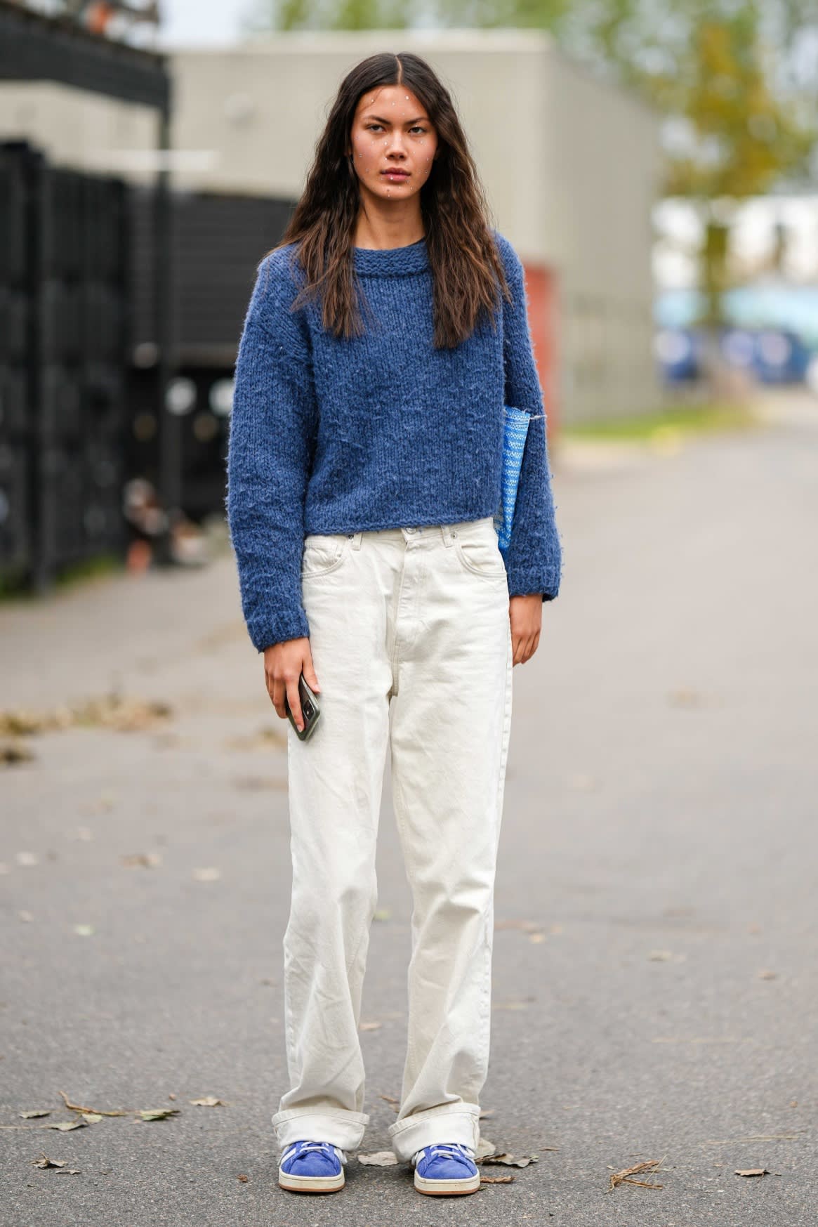 A woman stands in the street, wearing white trousers and blue sweater