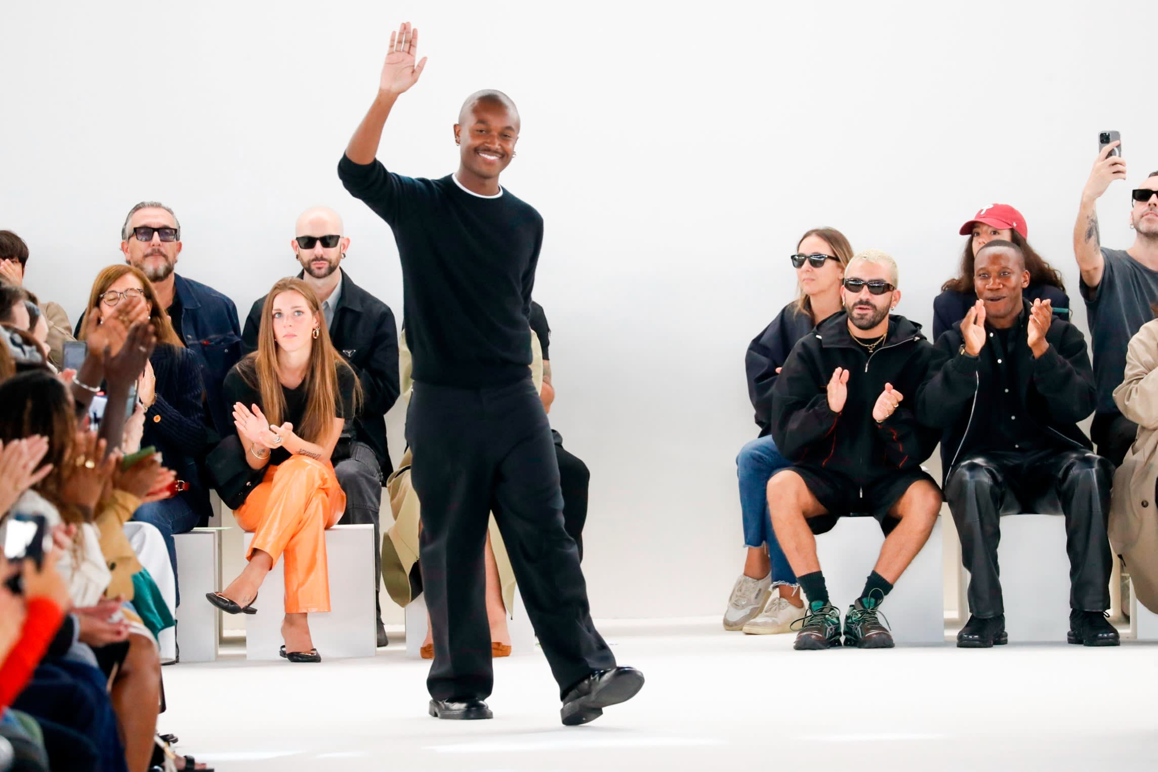 A young man in black T-shirt and trousers walks on a catwalk, smiling and waving