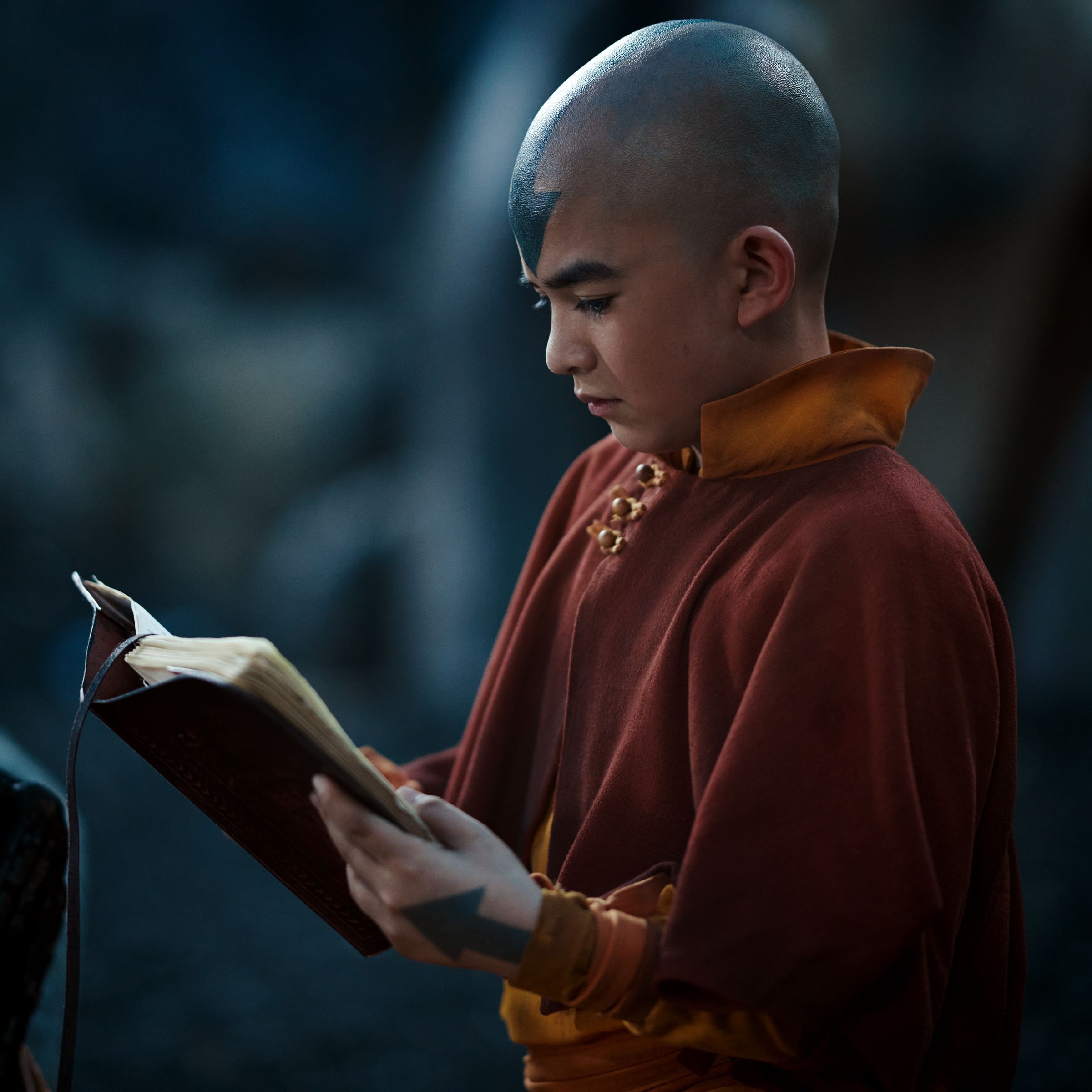 A bald boy with a blue arrow tattooed on his head. The boy is wearing an orange jumpsuit-like piece of clothing with a short amber cape around his neck. He’s also reading a book.