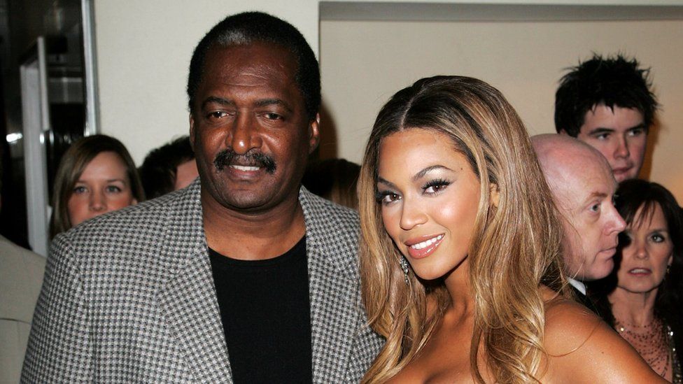 Dr Matthew Knowles pictured with Beyoncé at the Dream Girls in 2007. Dr Knowles, a black man in his 70s, has short black hair, brown eyes and a moustache. He wears a black and white dog-tooth blazer over a black top. Beyoncé is a black woman who in this picture would have been in her mid 20s. She has long honey-coloured hair and smiles at the camera. A crowd of people are pictured behind the pair