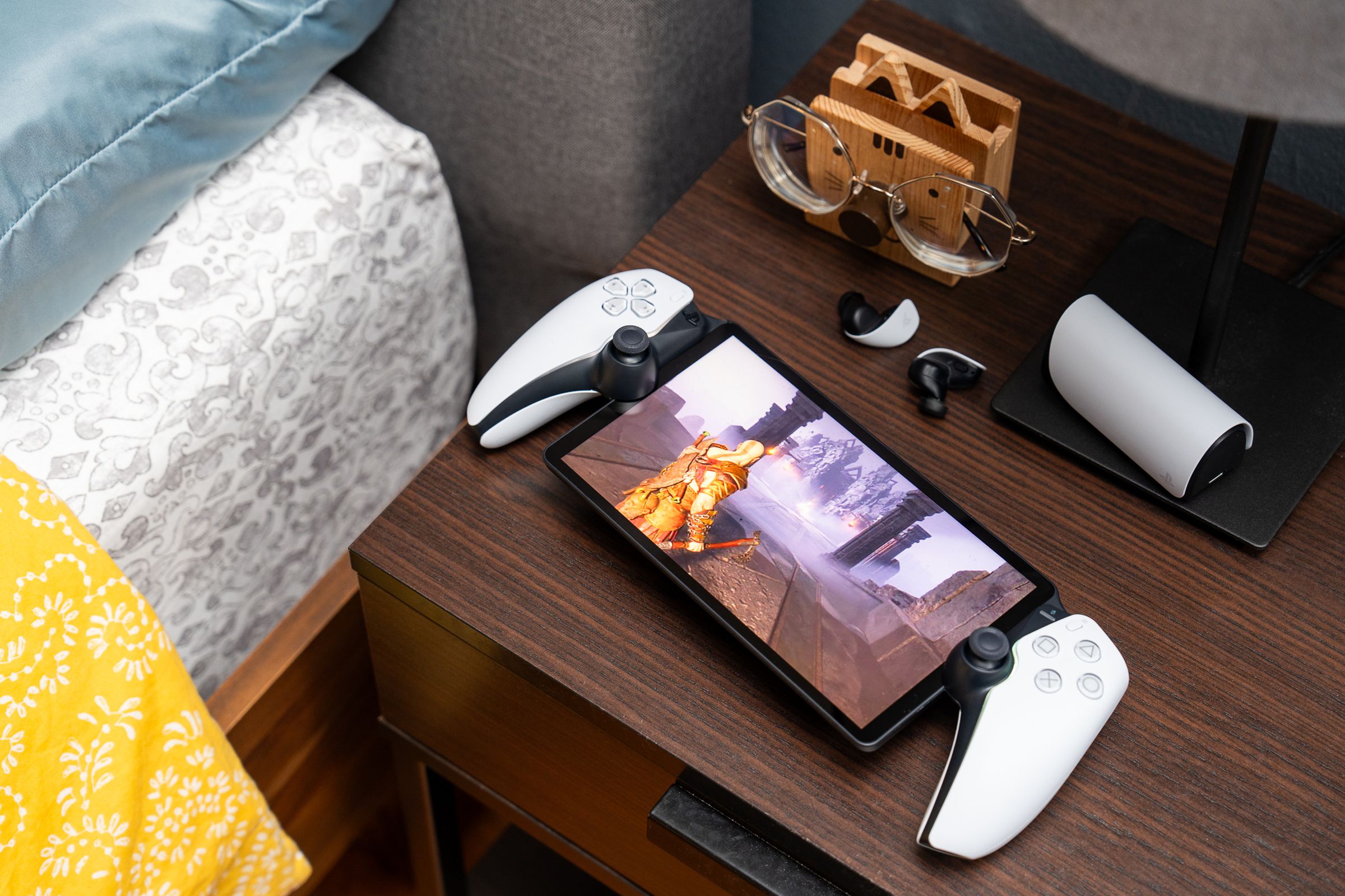 The PlayStation Portal sitting on a bedside table with a pair of earbuds. The handheld gaming device is streaming God of War: Ragnarök off a PlayStation 5.