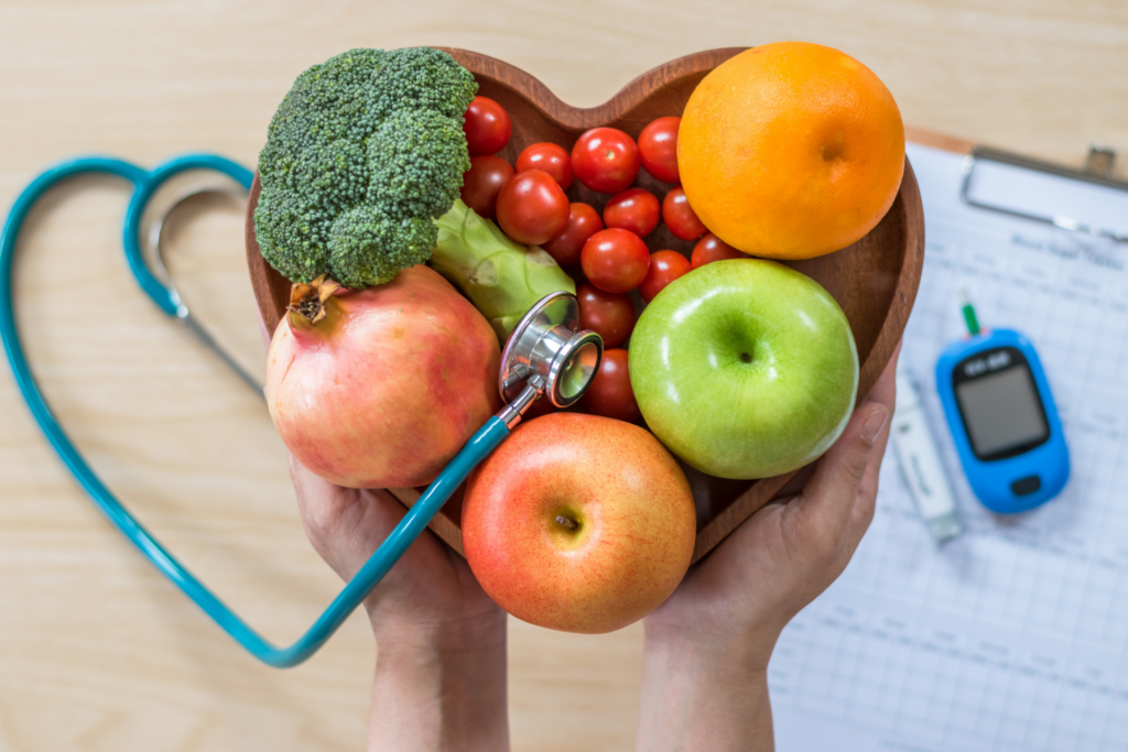 A wooden bowl in the shape of a heart holds fruits and vegetables. There is also a stethoscope.