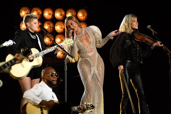 Beyoncé is holding a microphone onstage and is leaning toward a guitarist on her right. To her left, a fiddler smiles at the audience.