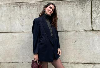 fashion editor wearing black blazer, black miniskirt, and gray turtleneck and standing in front of a brick wall