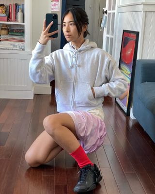 woman kneeling on wood floor in gray zip up hoodie sweatshirt with shorts and red socks and black sneakers taking selfie