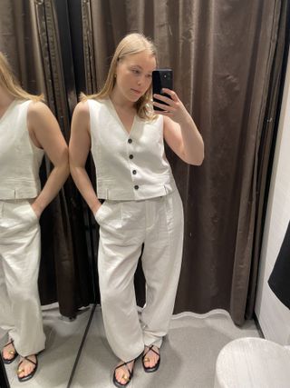 Woman in dressing room wears white linen waistcoat, linen trousers, sandals