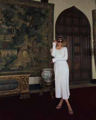 woman wearing white rib knit long sleeve midi dress with oversized straw sun hat, black sunglasses, black strappy sandals, in front of a wall tapestry