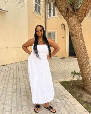 woman wearing white seamed linen dress with black thong flat sandals and pendant necklace and sunglasses standing on stone in front of old building