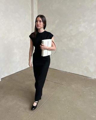 woman wearing black column dress with cap sleeve and black Mary Jane shoes, holding white structured shoulder bag, against marbled wall