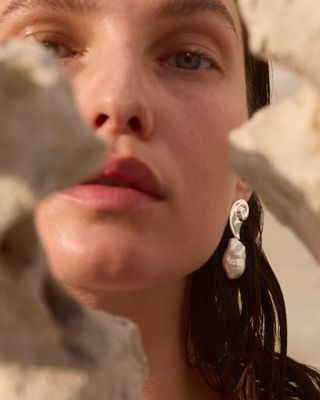 Woman peeking behind rock wearing silver shell pearl drop Jenny Bird earrings.