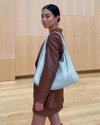 female fashion influencer Sasha Mei poses in an all-wood room wearing a low bun, earrings, light blue Bottega Veneta Intrecciato Hobo Bag, and brown skirt suit