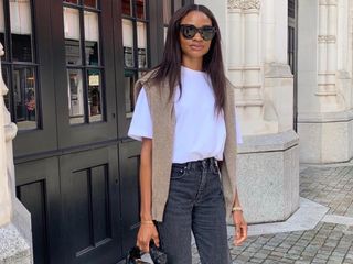 British female influencer Lorna Humphrey poses on the sidewalk in London wearing oversize black sunglasses, an oversize white T-shirt, tan sweater draped over the shoulders, and gray jeans