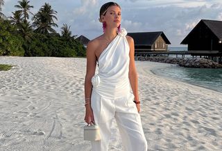 Sofia Richie Grainge wearing a white one-shoulder top, white trousers, and an off-white Loro Piana Extra Pocket top-handle vanity case bag on her honeymoon.