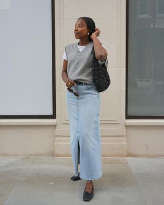 Woman wears grey vest, white t-shirt, denim skirt, black ballet flats and black COS bag