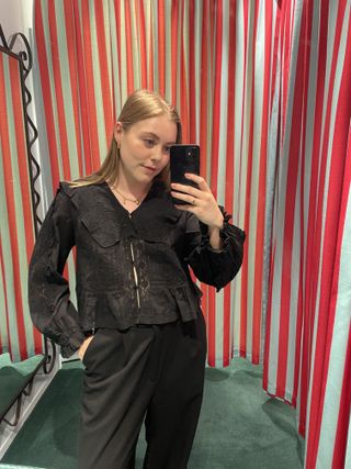 Woman in dressing room wears black blouse and black tailored trousers.