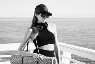 Kaia Gerber wearing a Celine hat, sunglasses, sports bra, bike shorts, large headphones, and a yoga mat hanging off her shoulder.