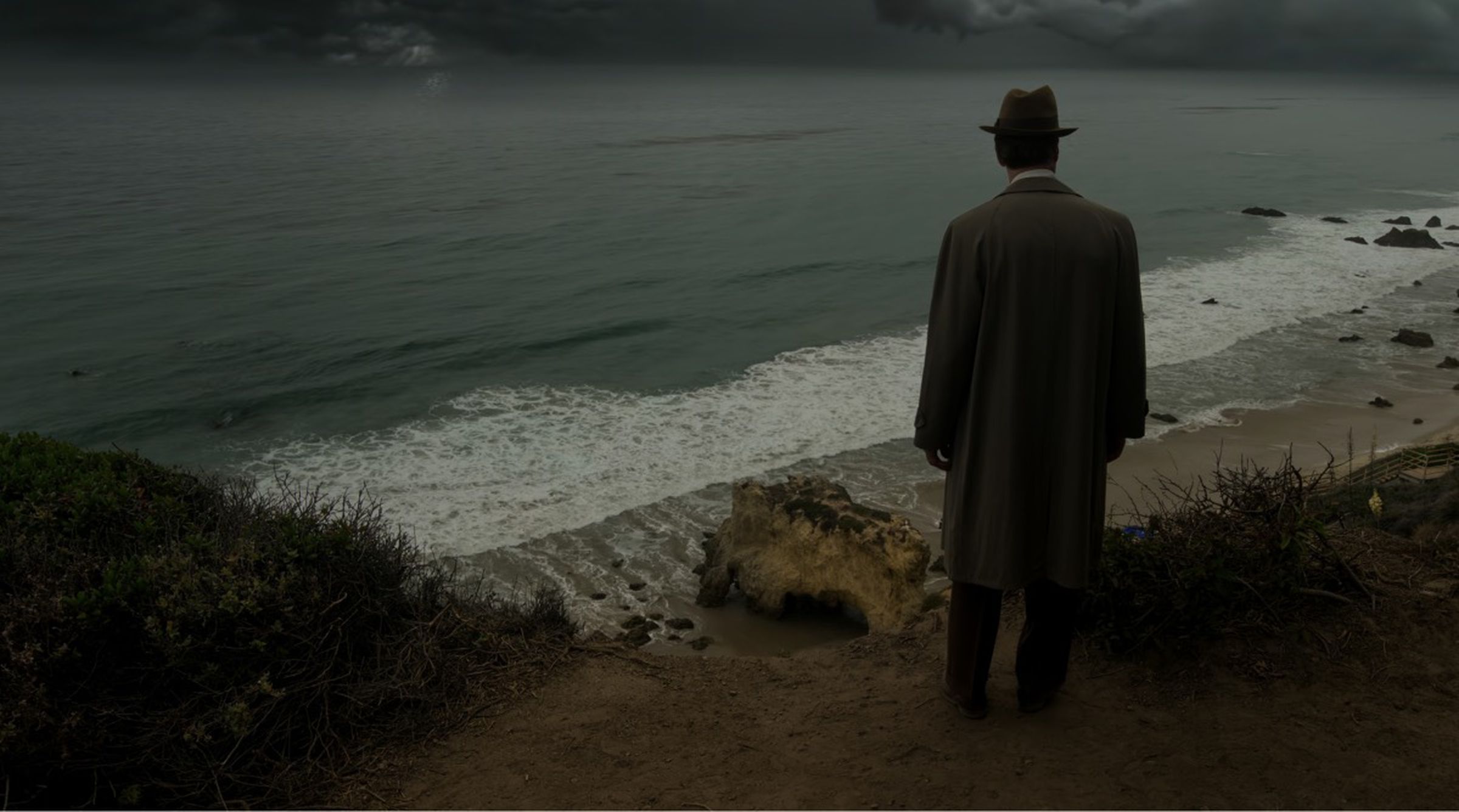 An image of a man in a fedora and coat standing on a cliff looking down at a stormy beach.