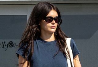 Kaia Gerber wearing sunglasses with a blue T-shirt and black skirt-over-pants look.