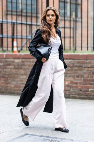 Katie Holmes wears white trousers, with a white tank top, mary janes, a black trench coat, and a black clutch.