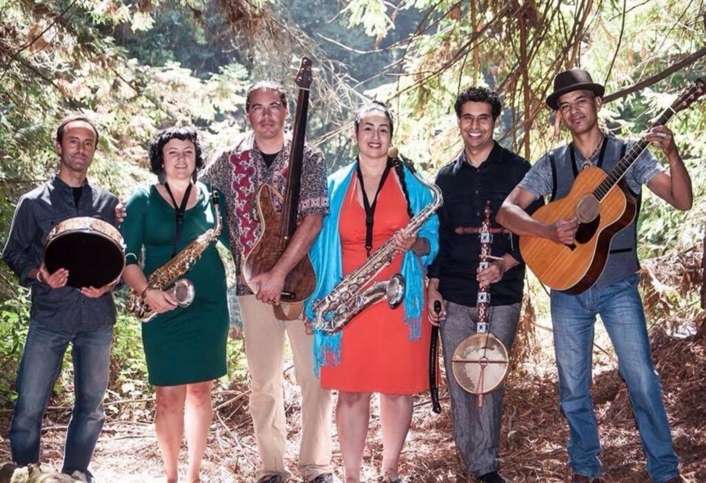 Alameda Post - A band of 6 people hold their instruments in the woods. 