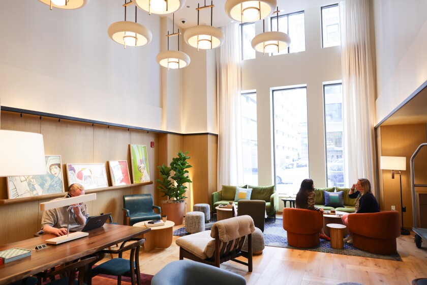 The lobby of L7 Chicago with some guests sitting in chairs talking by a window and another person sitting at a table working on a laptop.