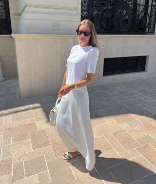 Swedish fashion influencer Lovisa Barkman walks outside on a neutral stone path wearing black cat-eye sunglasses, a white T-shirt, white maxi slip skirt, white top-handle bag, and white Hermès slide sandals.