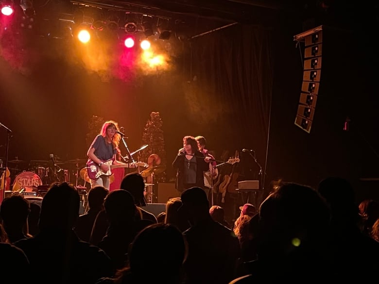 Two women sing into microphones on a dark stage in front of a rock band. A crowd is silhouetted in the foreground. The stage is misty and backlit