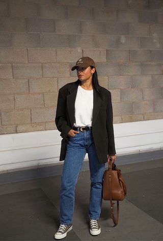 a photo of a summer rainy day outfit with a woman wearing a brown baseball cap with a black blazer, white t-shirt, straight-leg jeans, brown converse, and a brown handbag