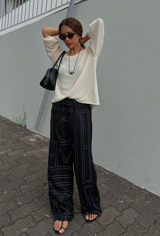 a photo of a summer rainy day outfit with a woman wearing a white lightweight sweater over a tank with printed silk trousers, black strappy flat sandals, a cord necklace, black sunglasses, and a black shoulder bag