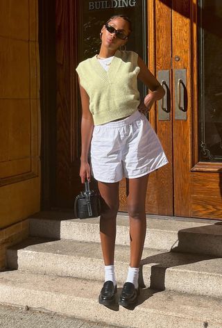 a photo of a summer rainy day outfit with a woman wearing a yellow sweater vest over a white t-shirt with white linen shorts, white socks, black loafers, black sunglasses, and a black handbag