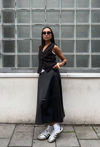 a photo of summer rainy day outfit with a woman wearing a black waistcoat over a white tank top with a black maxi skirt, white socks, samba sneakers, bangles, and black sunglasses