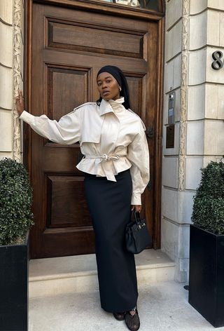 a photo of a summer rainy day outfit with a woman wearing a cropped tan trench coat over a black tank maxi dress with black fishnet mesh ballet flats, a black handbag, and a stretchy black headband