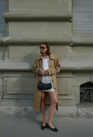 a photo of a summer rainy day outfit with a woman wearing a tan trench coat over a white mini dress with black ballet flats and a black shoulder bag