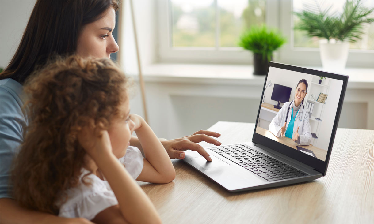 A parent sits with their child while they have a telehealth appointment on the laptop. A doctor is on the screen talking to them.